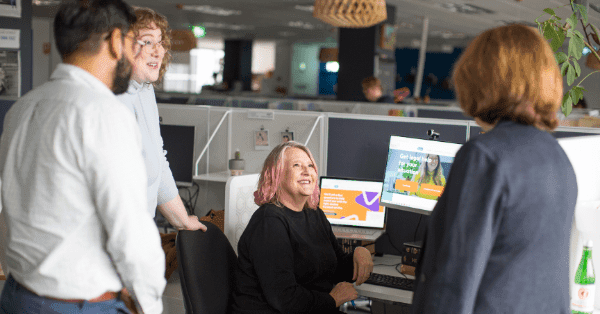 Four people are having a conversation in the office. One woman is sitting at a desk with computer screens, and the other three are standing around her