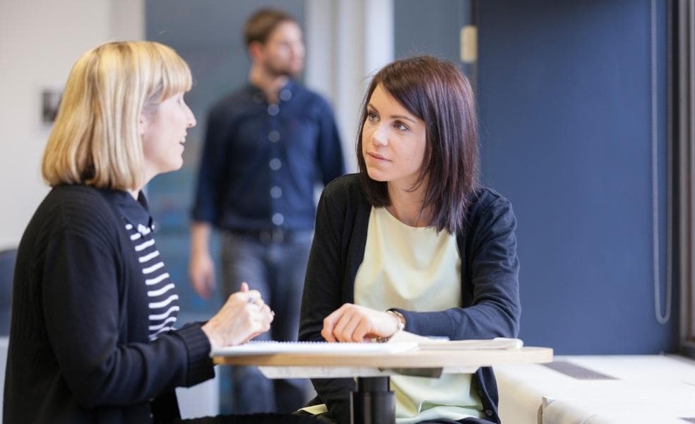 Two women talking. They are seated next to each other at a small table by an office window with their note pads