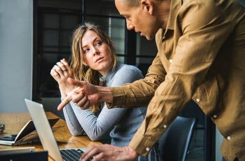 A man weraing brown shirt is explaining something to a woman sitting at a work table. He is pointing at a laptop screen.