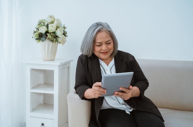 Older woman is sitting on a couch and reading off her tablet