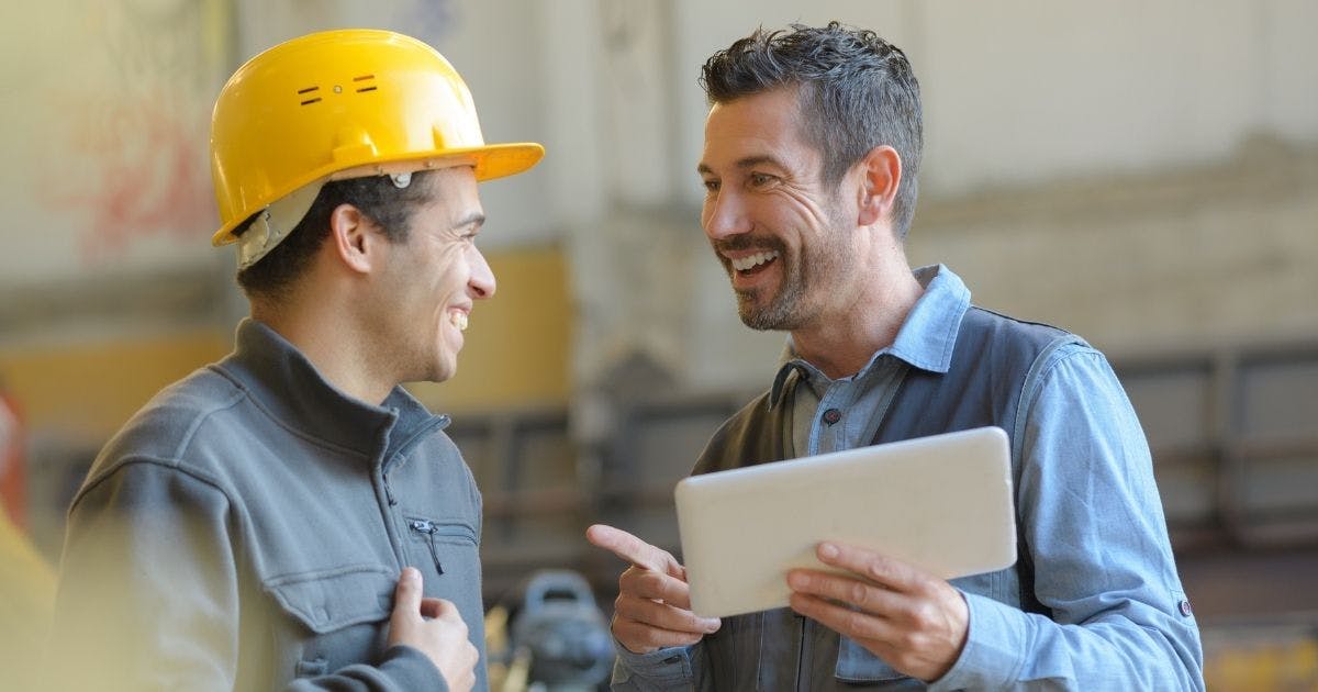 Photo of two men in a workplace, one wearing a hard hat