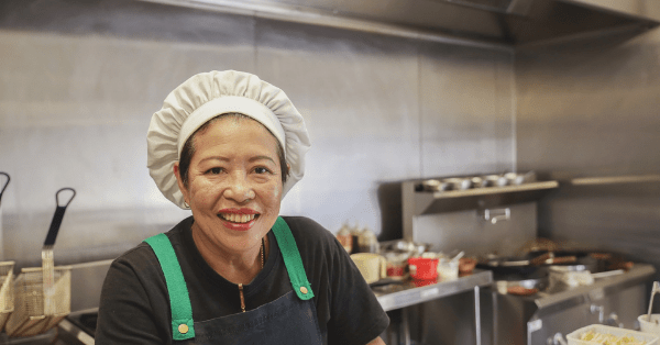Woman smiling at the camera. She is in a commercial kitchen wearing an apron and chefs hat