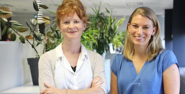 Two women in the office looking at the camera smiling with plants behind them