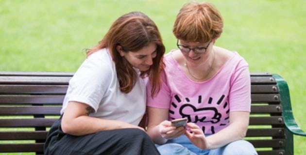 Two young people sitting on a park bench and looking at a mobile phone smiling