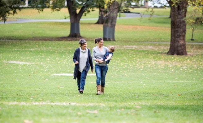 Two woman walking towards the camera through a park. One is holding a baby and they are both looking at the baby