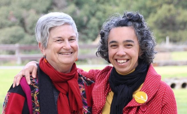 Two people smiling at the camera. They are both wearing red and colourful clothes, and one has their arm around the other.