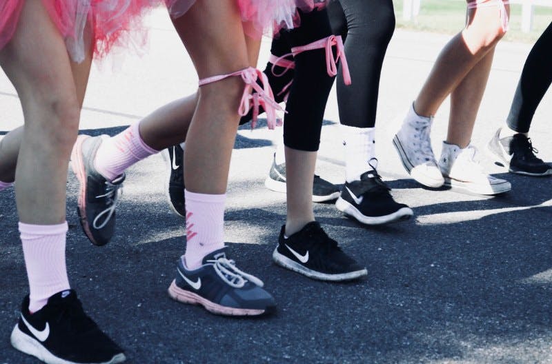 A group of people are in line about to run together. We can only see their legs and jogging shoes, and two people have pink ribbon tied around their knees.